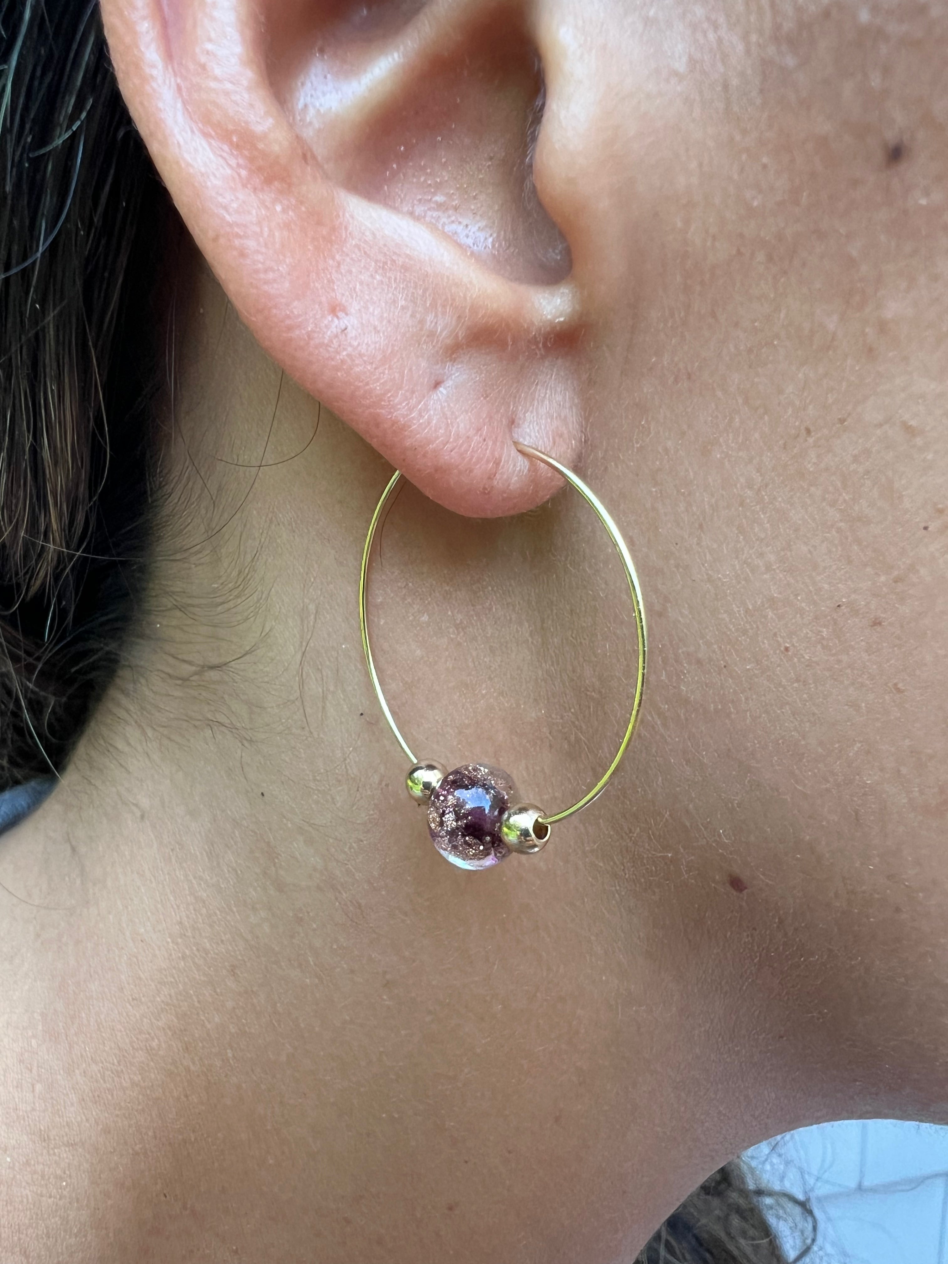 Gold hoop earring with a purple bead worn by a person.