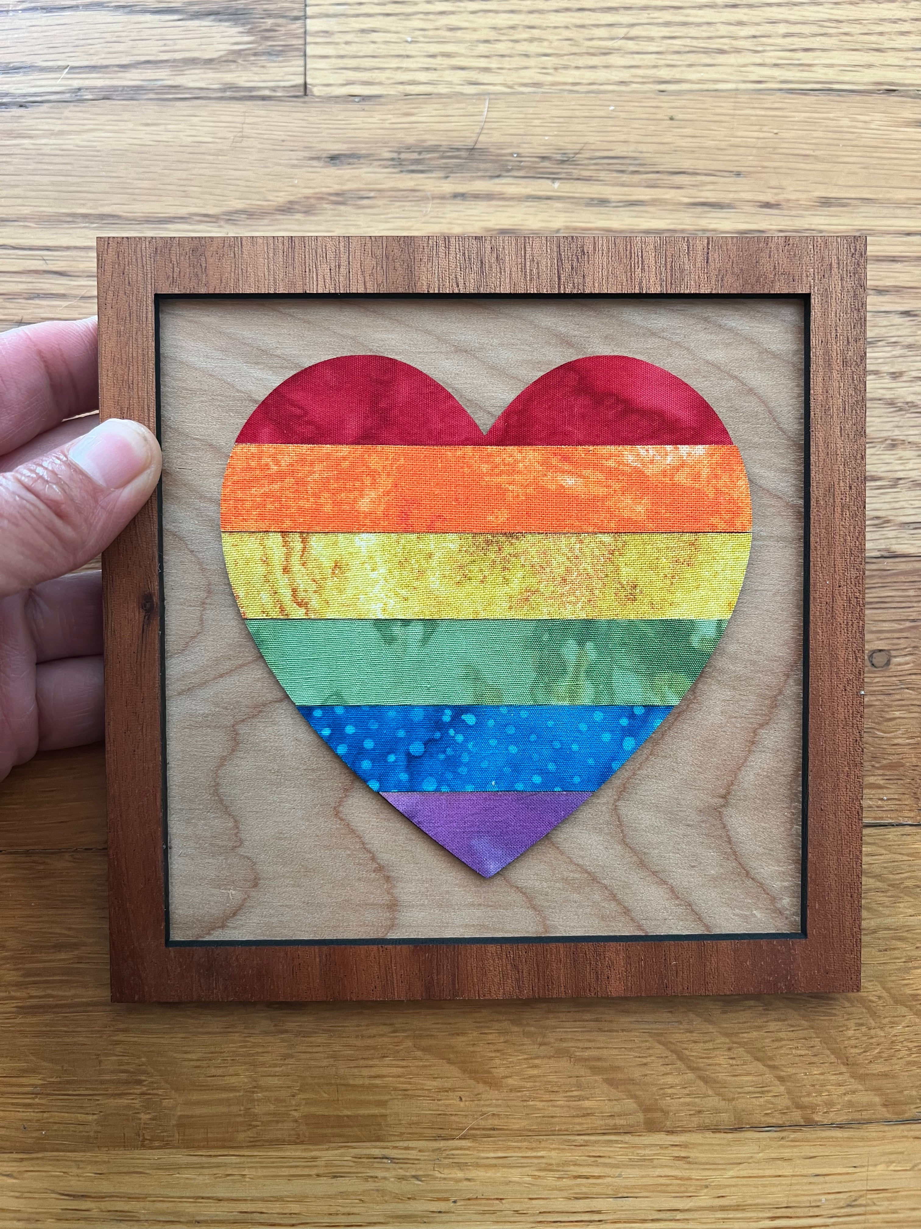 Rainbow heart-shaped artwork in a wooden frame held by a hand on a wooden surface