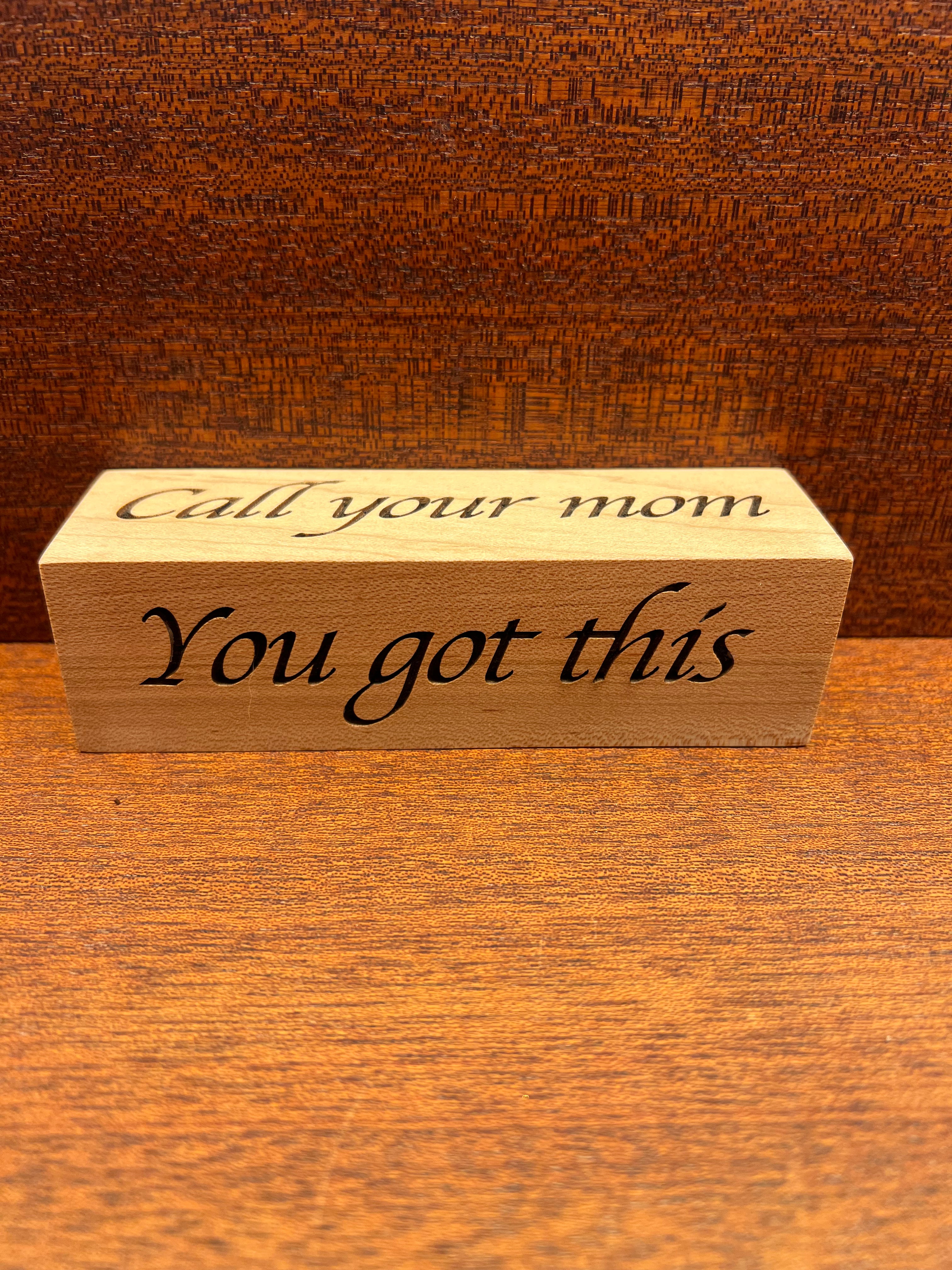 Wooden block with engraved text 'Call your mom You got this' on a wooden surface.