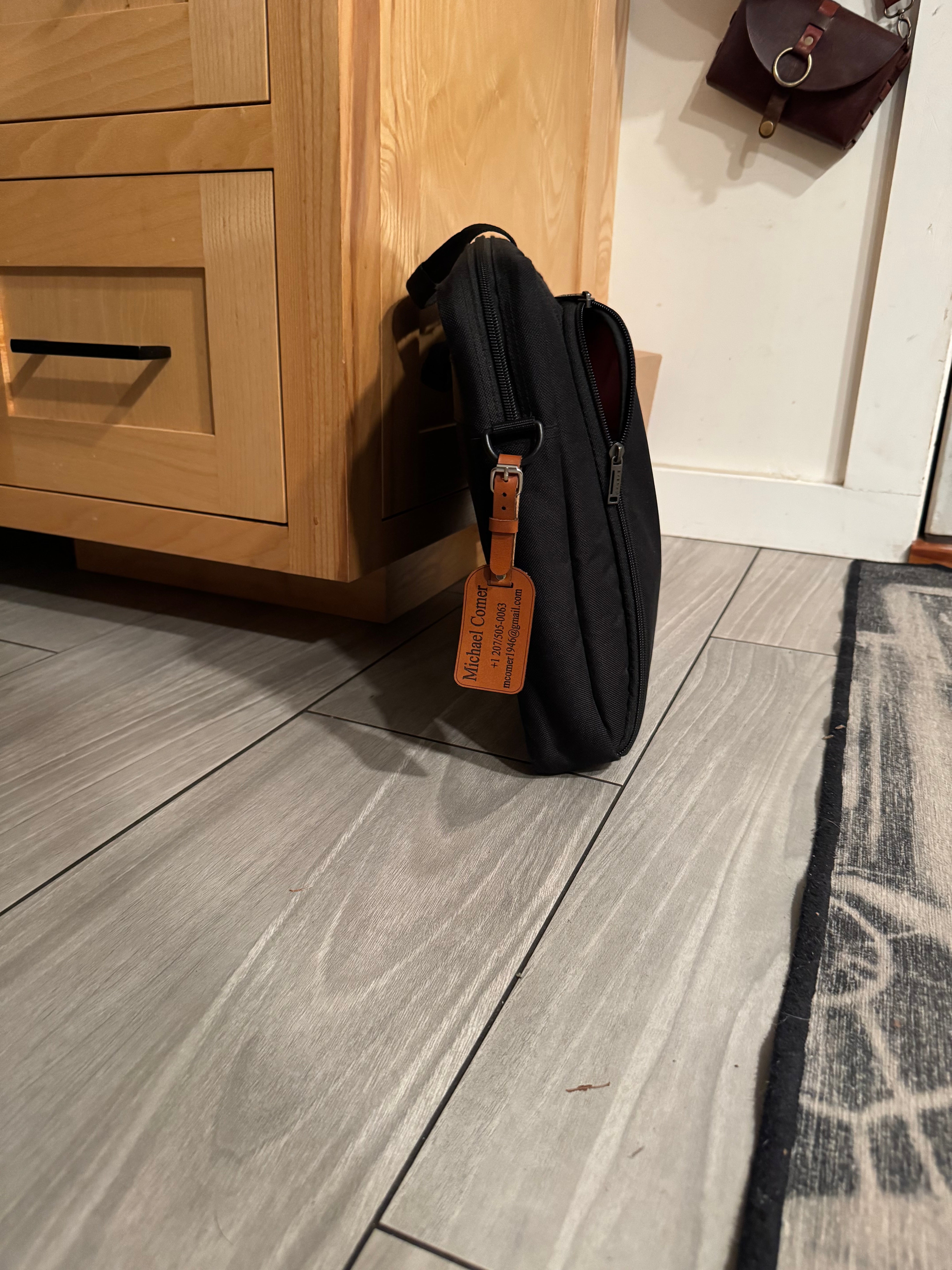 Black bag with a visible brand tag on a wooden surface against a gray tiled wall.